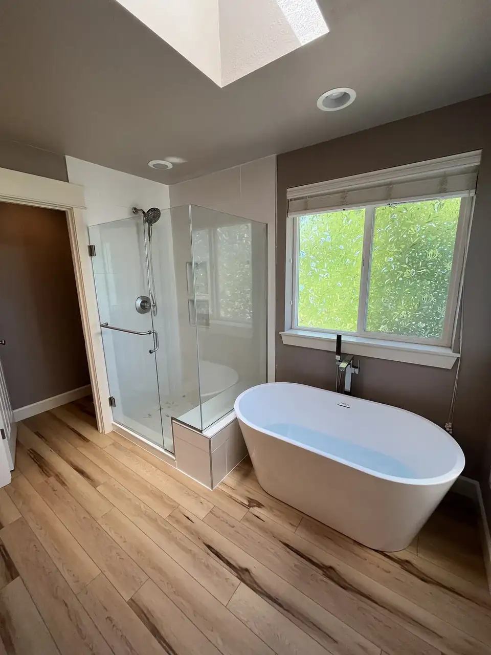 Bathroom remodeling Puyallup WA: primary bath with a freestanding soaking tub, frameless glass corner shower, skylight and wood-look plank floor