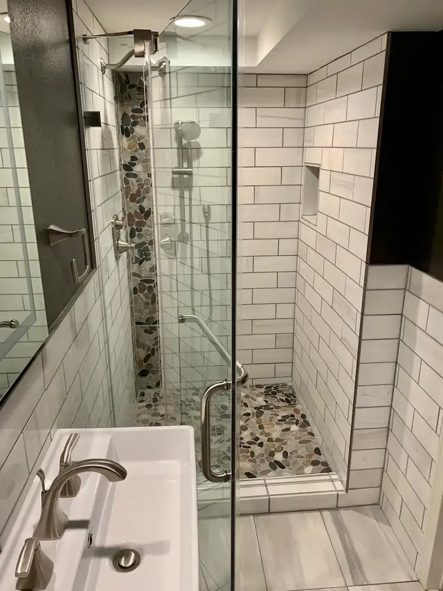 Subway tile walk-in shower with pebble accent strip and pebble floor behind frameless glass